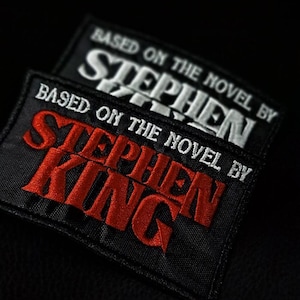 Può includere: Due toppe rettangolari nere con testo ricamato. La toppa superiore ha un testo bianco che recita "TRATTO DAL ROMANZO DI STEPHEN". La toppa inferiore ha un testo rosso che recita "TRATTO DAL ROMANZO DI STEPHEN KING".