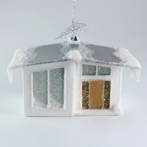 May include: A white, snow-covered, mid-century modern house ornament with a silver roof and a brown door. The house has a small antenna on top and icicle-like details on the eaves.