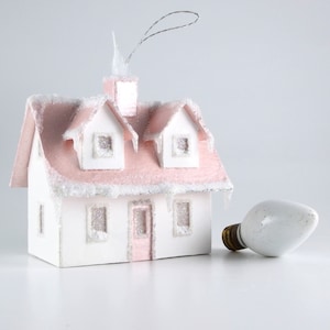 May include: A pink and white paper mache Christmas ornament shaped like a house with a light bulb. The house has a pink roof with white trim and windows. The house is decorated with glitter.