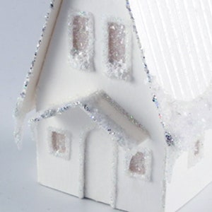 May include: White papercraft house with a snowy roof and glitter accents. The house has three windows and a front door.