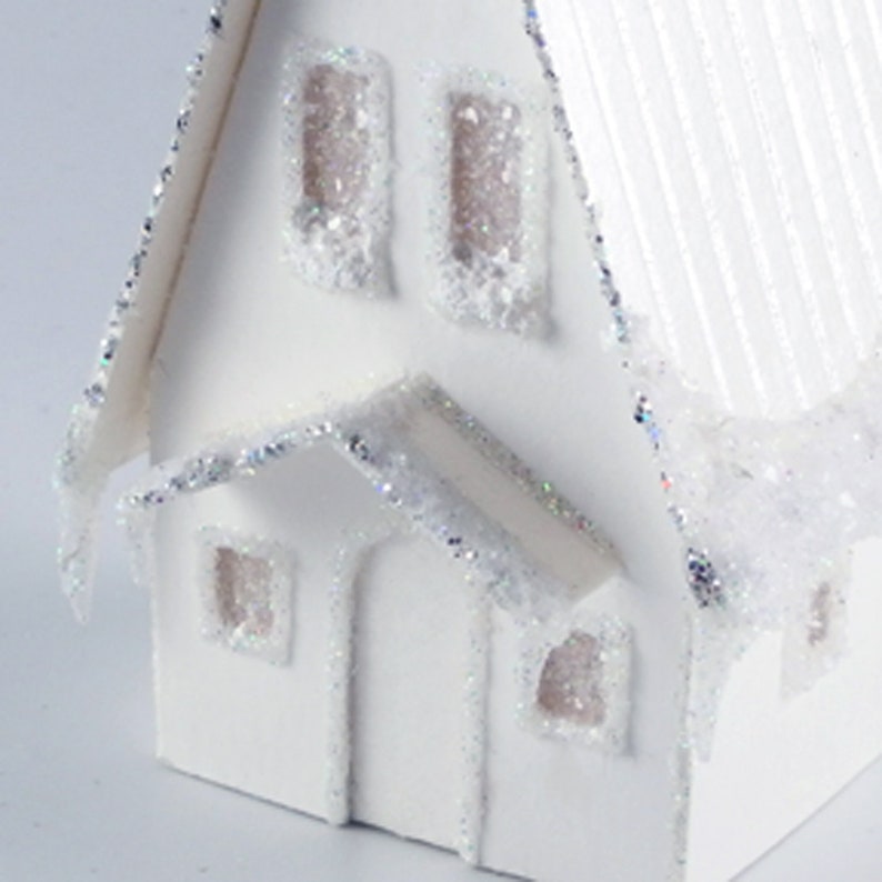 May include: White papercraft house with a snowy roof and glitter accents. The house has three windows and a front door.
