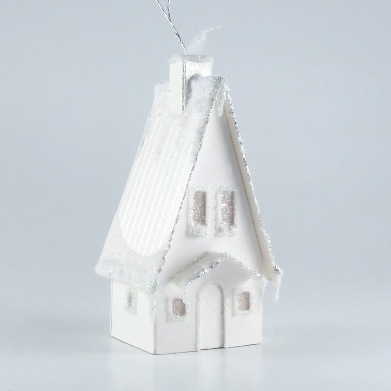 May include: White paper mache Christmas ornament shaped like a house with a snowy roof and icicle details. The house has a chimney, windows, and a door.