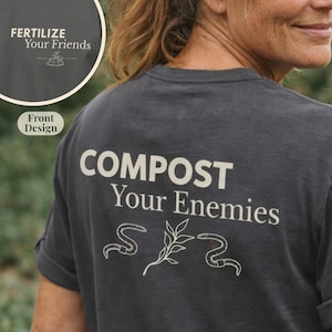 May include: Dark gray t-shirt with the text "COMPOST Your Enemies" in white block letters. The design includes illustrations of worms and a plant. The front of the shirt has the text "FERTILIZE Your Friends".