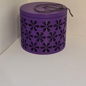 May include: A cylindrical purple container with a lid and a decorative floral pattern. The container has a dark gray string tied around the lid. The floral cutouts reveal a dark interior.