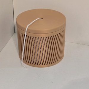 May include: A tan cylindrical container with a matching lid. The container has vertical, curved slots around the sides. A white string extends from a hole in the lid. The container is sitting on a white surface.