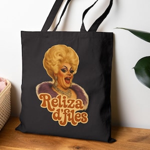 Könnte beinhalten: Schwarze Tragetasche mit einer Illustration im Vintage-Stil einer Dragqueen mit einer großen blonden Perücke, einem lila Pelzstola und einer goldenen Halskette. Der Text "Reliza d'Files" ist in einer Retro-Schriftart.