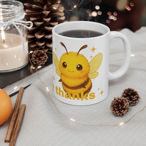 Puede incluir: Taza de cerámica blanca con una abeja de dibujos animados con pelaje amarillo y marrón, alas y el texto "thanks". La taza contiene un líquido oscuro y tiene un asa curva. Decoración con piñas y una vela.