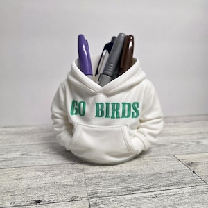 May include: A white miniature hoodie pen holder with the text "GO BIRDS" in green. The hoodie is filled with pens of various colors, including purple, gray, and brown. The pen holder sits on a light gray wooden surface.