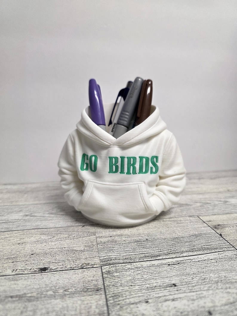 May include: A white miniature hoodie pen holder with the text "GO BIRDS" in green. The hoodie is filled with pens of various colors, including purple, gray, and brown. The pen holder sits on a light gray wooden surface.
