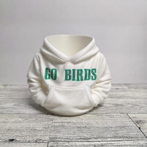 May include: A miniature white hoodie with the text "GO BIRDS" in green lettering across the chest. The hoodie features a hood and a front pocket, and is displayed on a light gray wooden surface.