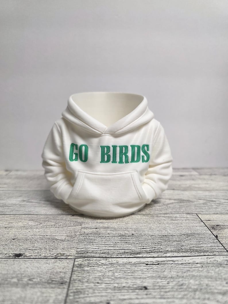 May include: A miniature white hoodie with the text "GO BIRDS" in green lettering across the chest. The hoodie features a hood and a front pocket, and is displayed on a light gray wooden surface.