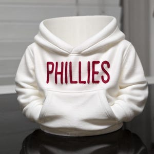 May include: White hooded sweatshirt with a kangaroo pocket. The word "PHILLIES" is printed in red across the chest. The sweatshirt is made of a soft material and has a hood.