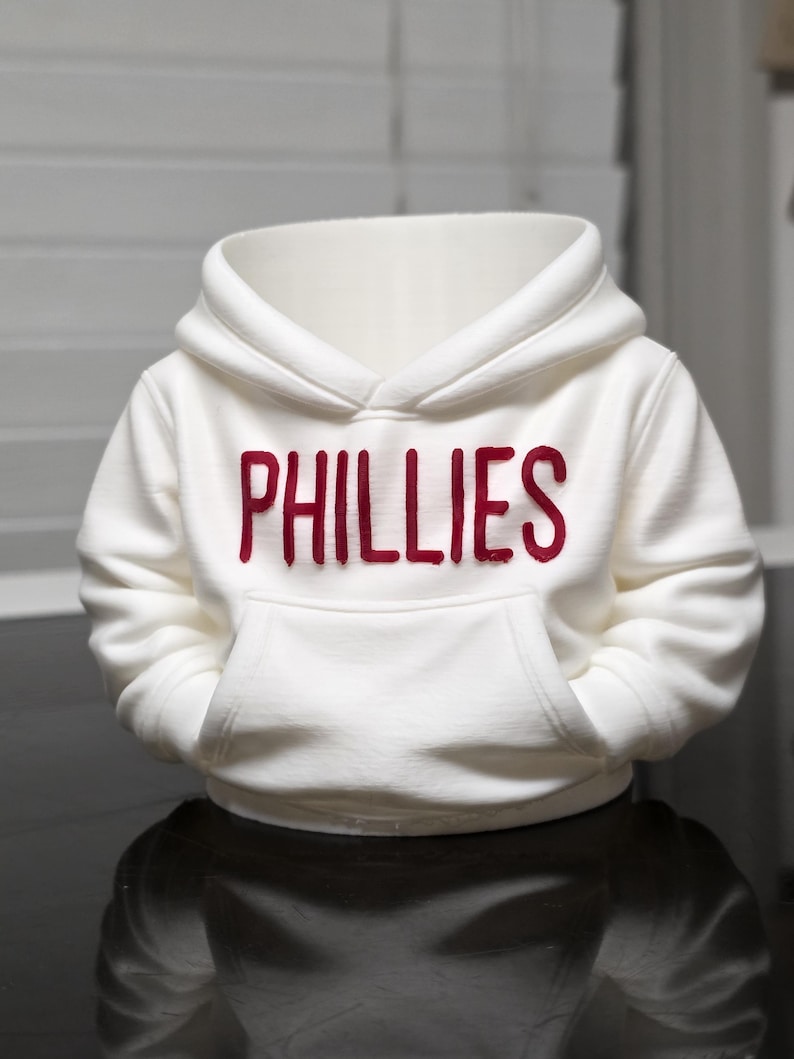 May include: White hooded sweatshirt with a kangaroo pocket. The word "PHILLIES" is printed in red across the chest. The sweatshirt is made of a soft material and has a hood.