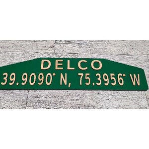 Peut inclure: Un panneau en bois vert avec le mot "DELCO" en lettres marron clair. En dessous, les coordonnées "39.9090° N, 75.3956° W" sont inscrites dans la même couleur marron clair. Le panneau est rectangulaire avec une extrémité pointue.