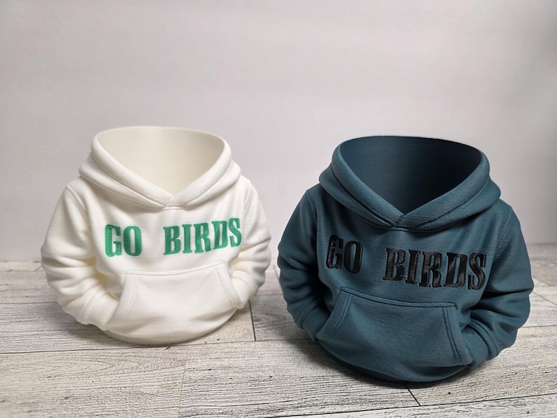 May include: Two miniature hoodie-shaped planters. One is white with green embroidered text reading "GO BIRDS", the other is teal with black embroidered text. Both have a hood and a front pocket.
