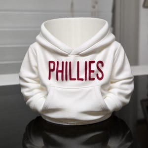 May include: A white hooded sweatshirt with the word "PHILLIES" in red block letters across the chest. The sweatshirt has a kangaroo pocket and a drawstring hood. The garment is displayed on a reflective surface.