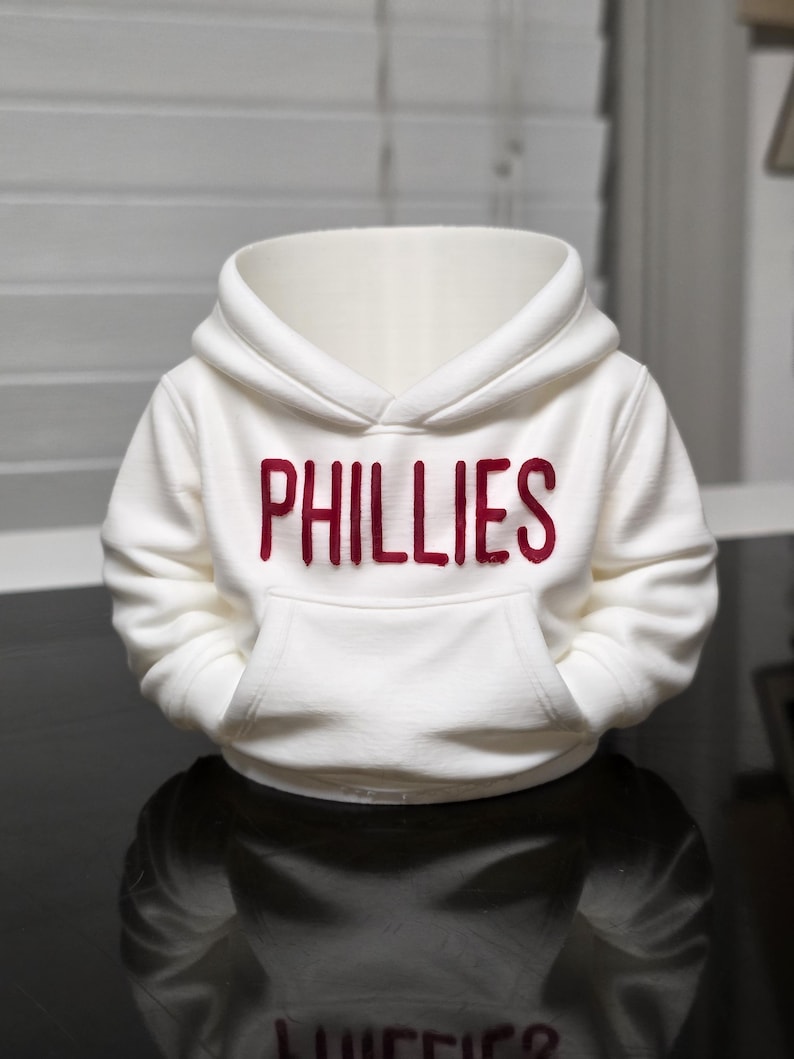 May include: A white hooded sweatshirt with the word "PHILLIES" in red block letters across the chest. The sweatshirt has a kangaroo pocket and a drawstring hood. The garment is displayed on a reflective surface.