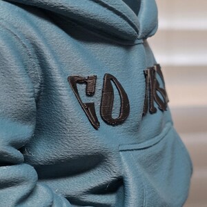 May include: A teal hoodie with a hood and a front pocket. The word "FOR" is printed in black on the upper back of the hoodie. The fabric has a textured appearance.