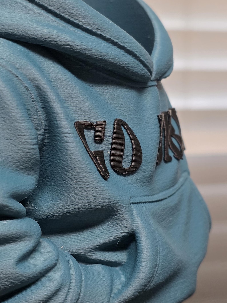 May include: A teal hoodie with a hood and a front pocket. The word "FOR" is printed in black on the upper back of the hoodie. The fabric has a textured appearance.