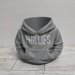 May include: A gray hoodie with the word "PHILLIES" printed in white across the chest. The hoodie has a front pocket and a hood. The item is sitting on a light gray and white wooden surface.