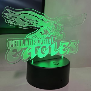 May include: A glowing green Philadelphia Eagles logo night light. The logo features an eagle with outstretched wings above the words "Philadelphia Eagles". The light sits on a black circular base.