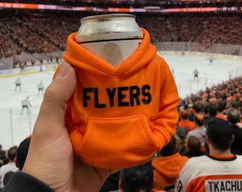 May include: A bright orange miniature hoodie beverage insulator featuring the word FLYERS in bold black lettering across the chest. The hoodie includes a hood, long sleeves, and a front pouch pocket, designed to fit snugly around a standard drink can.