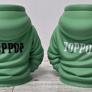 May include: Two green hoodie-shaped planters. One has "POPPDP" in black lettering on the back, and the other has "YOPPOP" in white lettering. The planters are on a light-colored surface.