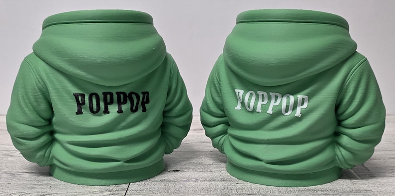 May include: Two green hoodie-shaped planters. One has "POPPDP" in black lettering on the back, and the other has "YOPPOP" in white lettering. The planters are on a light-colored surface.