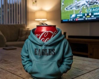 May include: A teal-colored miniature hoodie beverage insulator featuring the word EAGLES printed in black across the chest. The fabric garment is designed to fit around a standard red and white Coca-Cola aluminum soda can.