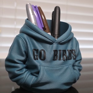 May include: A teal pen holder shaped like a hoodie, with the words "GO BIRDS" in black lettering. The pen holder has a front pocket and contains several pens in silver, purple, brown, and black.