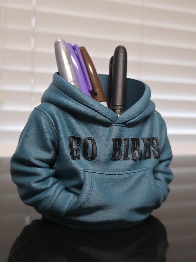 May include: A teal pen holder shaped like a hoodie, with the words "GO BIRDS" in black lettering. The pen holder has a front pocket and contains several pens in silver, purple, brown, and black.