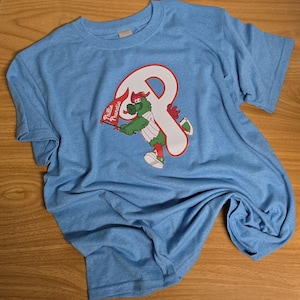May include: Light blue t-shirt featuring a graphic of the Philadelphia Phillies mascot, a green creature with a red and white baseball uniform, and the text "Phillie Phanatic". The mascot is holding a flag. The shirt is on a wooden surface.