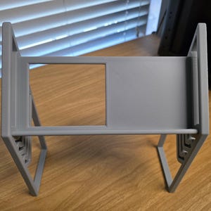 May include: A gray, 3D-printed stand with a rectangular frame and angled legs. The stand features a large rectangular opening in the center and a textured surface. The design is modern and minimalist, suitable for holding various items.