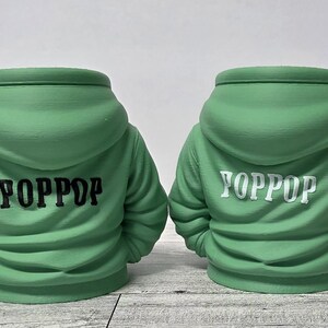May include: Two green hoodie-shaped planters. The back of each planter is visible, with the word "YOPPOP" printed on the back. One planter has black text, and the other has white text. The planters are on a light-colored surface.