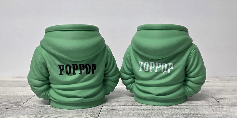 May include: Two green hoodie-shaped planters. The back of each planter is visible, with the word "YOPPOP" printed on the back. One planter has black text, and the other has white text. The planters are on a light-colored surface.