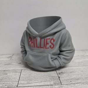 May include: A gray hoodie with the word "PHILLIES" in red letters across the chest. The hoodie has a front pocket and a hood. The item is a decorative object, possibly a planter or a unique storage container.