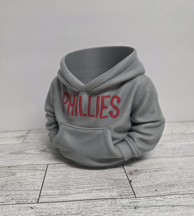 May include: A gray hoodie with the word "PHILLIES" in red letters across the chest. The hoodie has a front pocket and a hood. The item is a decorative object, possibly a planter or a unique storage container.