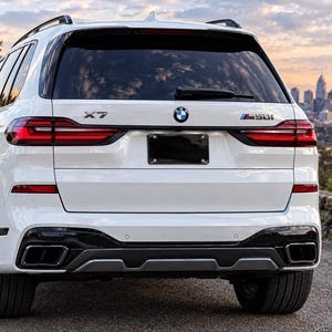 May include: A white BMW X7 M50i SUV viewed from the rear. The vehicle features a black number plate frame, dark tinted windows, a rear windscreen wiper, and dual exhaust outlets integrated into a black and grey lower bumper trim.