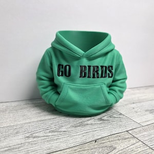 May include: A green hoodie with the text "GO BIRDS" in black lettering. The hoodie has a front pocket and a hood. The garment is displayed on a white background and a wooden surface.
