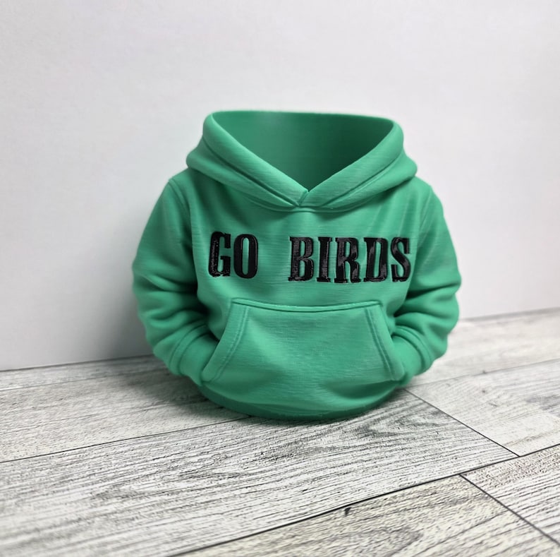 May include: A green hoodie with the text "GO BIRDS" in black lettering. The hoodie has a front pocket and a hood. The garment is displayed on a white background and a wooden surface.