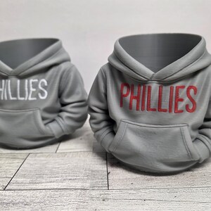May include: Two gray hoodie-shaped planters with the word "PHILLIES" across the front. One has white lettering, the other red. The planters have a hood and a front pocket, resembling miniature sweatshirts. They sit on a light wood-grain surface.