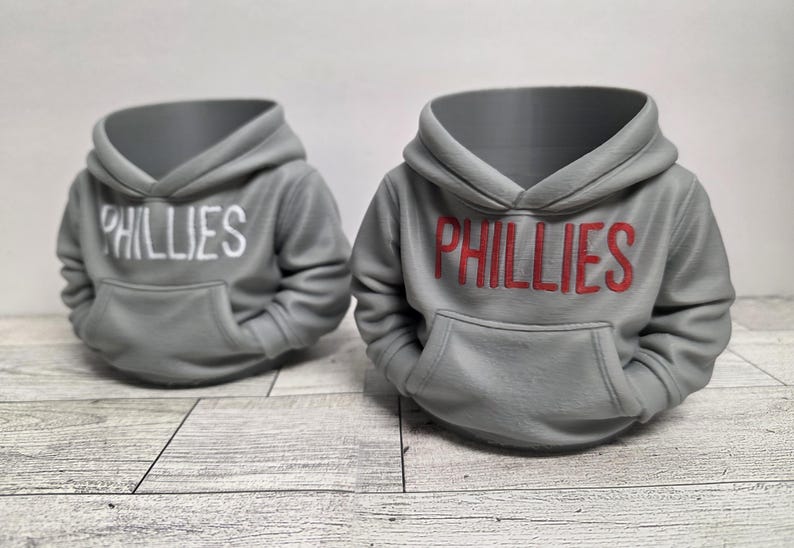 May include: Two gray hoodie-shaped planters with the word "PHILLIES" across the front. One has white lettering, the other red. The planters have a hood and a front pocket, resembling miniature sweatshirts. They sit on a light wood-grain surface.