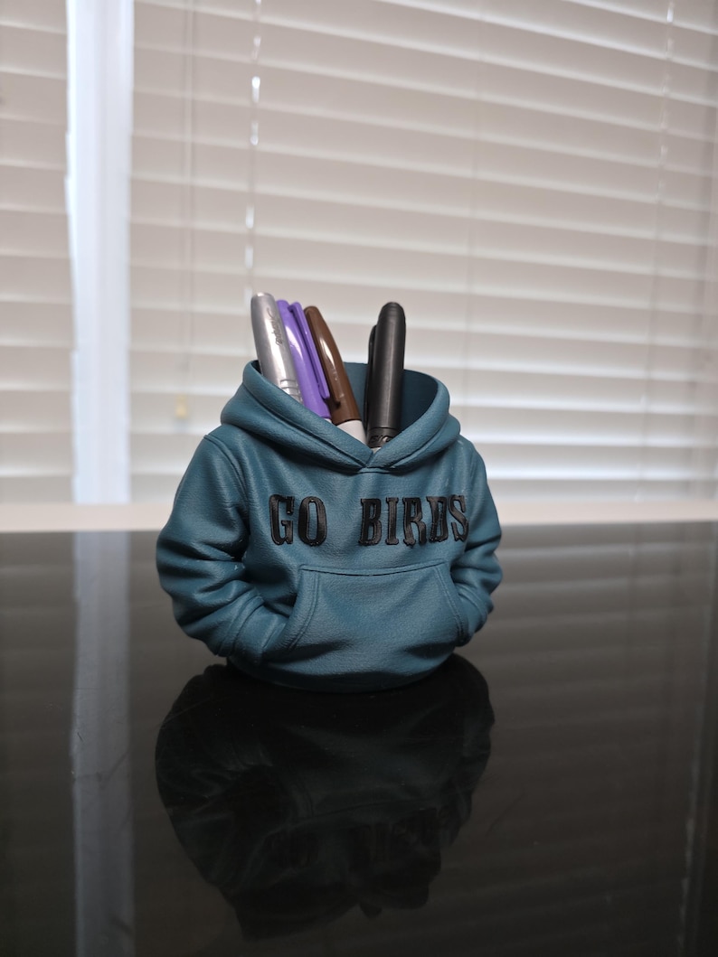 May include: A teal-colored pen holder shaped like a hoodie, with the words "GO BIRDS" across the front. The pen holder contains several pens in silver, purple, brown, and black. The pen holder is sitting on a reflective surface.