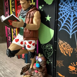May include: A figurine of a muscular character wearing a brown vest, green shirt, and white shorts with red hearts, reading a book. A small, furry creature with purple glasses and a drink sits at the character's feet against a black backdrop.