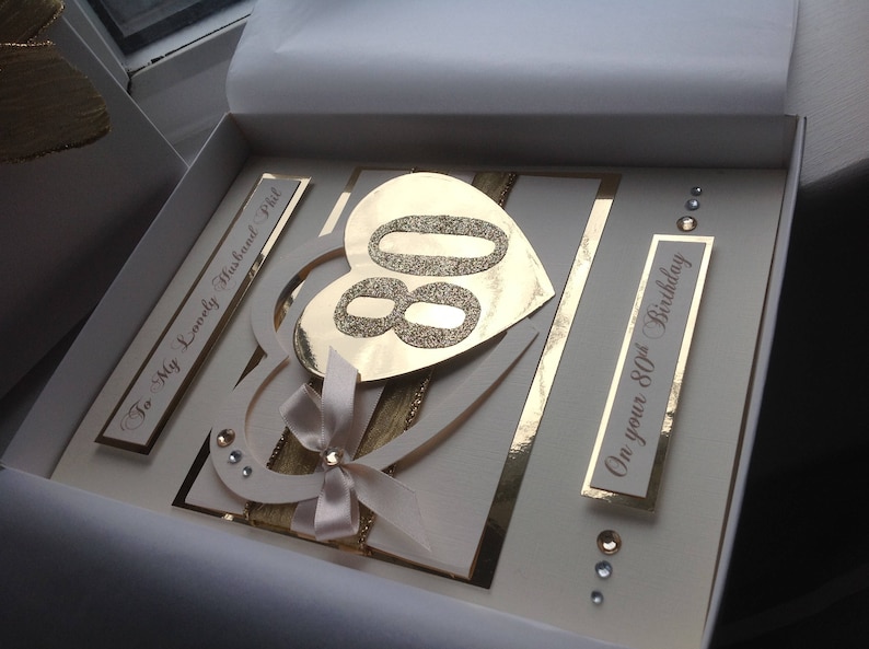 May include: A white card with gold accents and a gold glitter heart with the number 80 inside. The card says "To my Lovely Husband Phil" and "On your 80th Birthday".