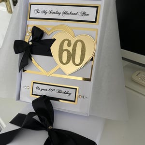 May include: A white card with a black ribbon and gold glitter hearts. The card says "To My Darling Husband Alan" and "On your 60th Birthday".