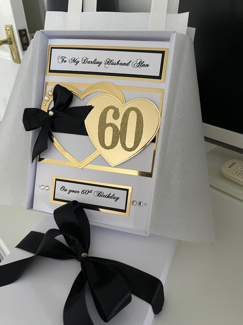 May include: A white card with a black ribbon and gold glitter hearts. The card says "To My Darling Husband Alan" and "On your 60th Birthday".