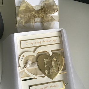 May include: A white card with a gold glitter heart with the number 50 on it. The card is in a white box with a gold ribbon. The card says "To My Lovely Husband Jeff" and "On your 50th Birthday".