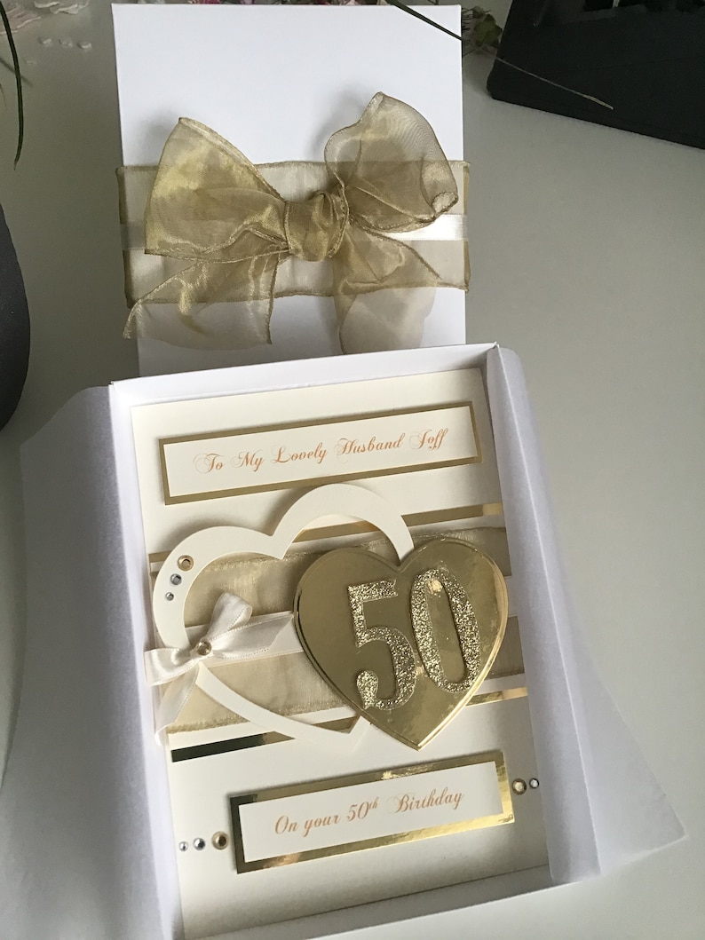 May include: A white card with a gold glitter heart with the number 50 on it. The card is in a white box with a gold ribbon. The card says "To My Lovely Husband Jeff" and "On your 50th Birthday".