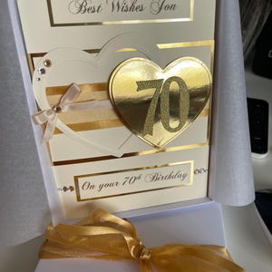 May include: A white card with gold accents and a gold heart with the number 70 on it. The card says "Best Wishes Son" and "On your 70th Birthday".